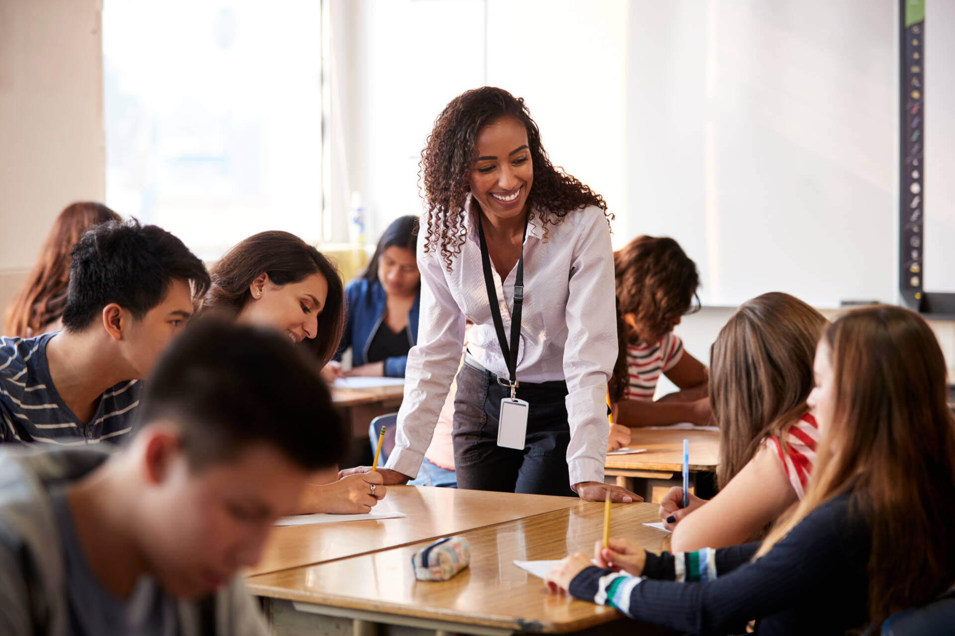 Female High School Teacher Standing By Student Table Teaching Lesson Female High School Teacher Standing By Student Table Teaching Lesson